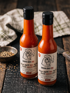 Two bottles of Smokin Bros BBQ Good Eats Red Sauce (Sauce Rouge EATS) standing on a rustic dark wooden table with a crumpled white cloth and a small bowl of peppercorns in the background.
