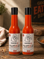 Two bottles of Smokin Bros BBQ Good Eats Red Sauce standing on a rustic wooden surface, with a blurred warm-toned background and a sign reading Good Eats BBQ.