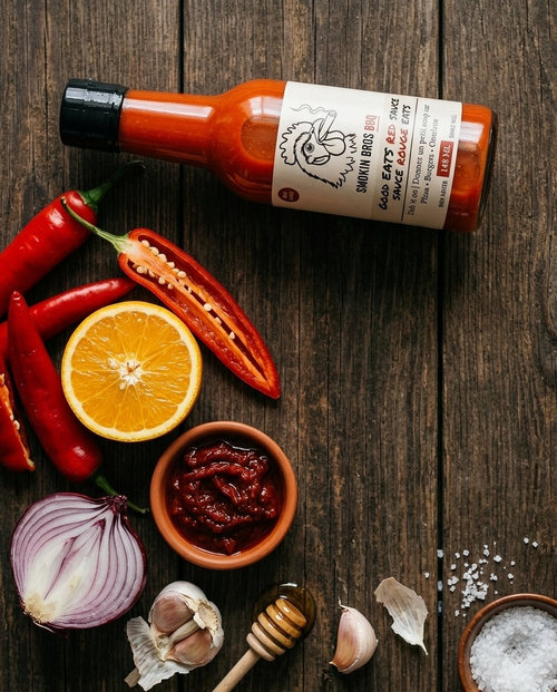 A bottle of Smokin Bros BBQ Good EATS Red sauce lies on a dark wooden tabletop surrounded by food ingredients including red chili peppers, a sliced orange, a small bowl of red sauce, garlic cloves, an onion slice, a honey dipper, and coarse salt.