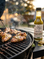 A bottle of Smokin' Bros BBQ Good EATS Green Sauce next to a black grill with chicken wings cooking, set in an outdoor backyard environment. A brush with green sauce is next to the bottle.