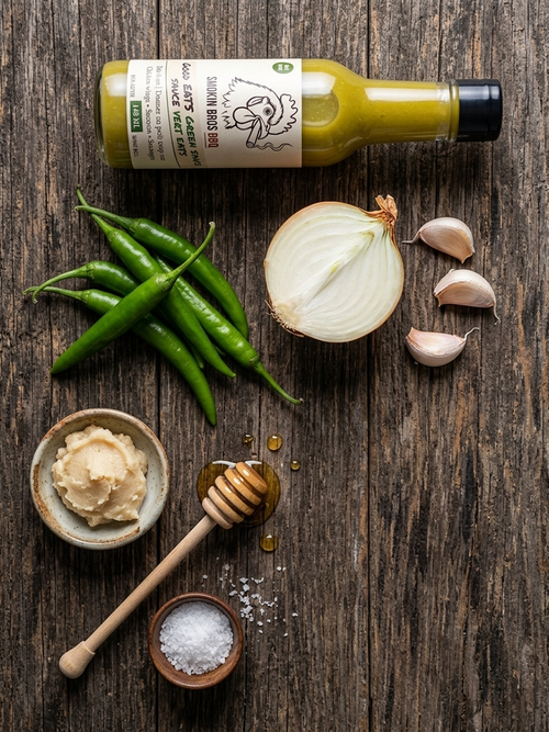 A bottle of Smokin Bros BBQ Good EATS Green Sauce placed on a rustic dark wood surface, surrounded by fresh ingredients including green chilies, a halved white onion, garlic cloves, a small bowl of mustard, a honey dipper, and a small bowl of sea salt.