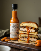 A bottle of Smokin Bros BBQ Sweet Heat Habanero Sauce stands next to a stacked grilled chicken sandwich on a wooden board, with sauce dripping from the top. Hot peppers and additional sauce are scattered on the board.
