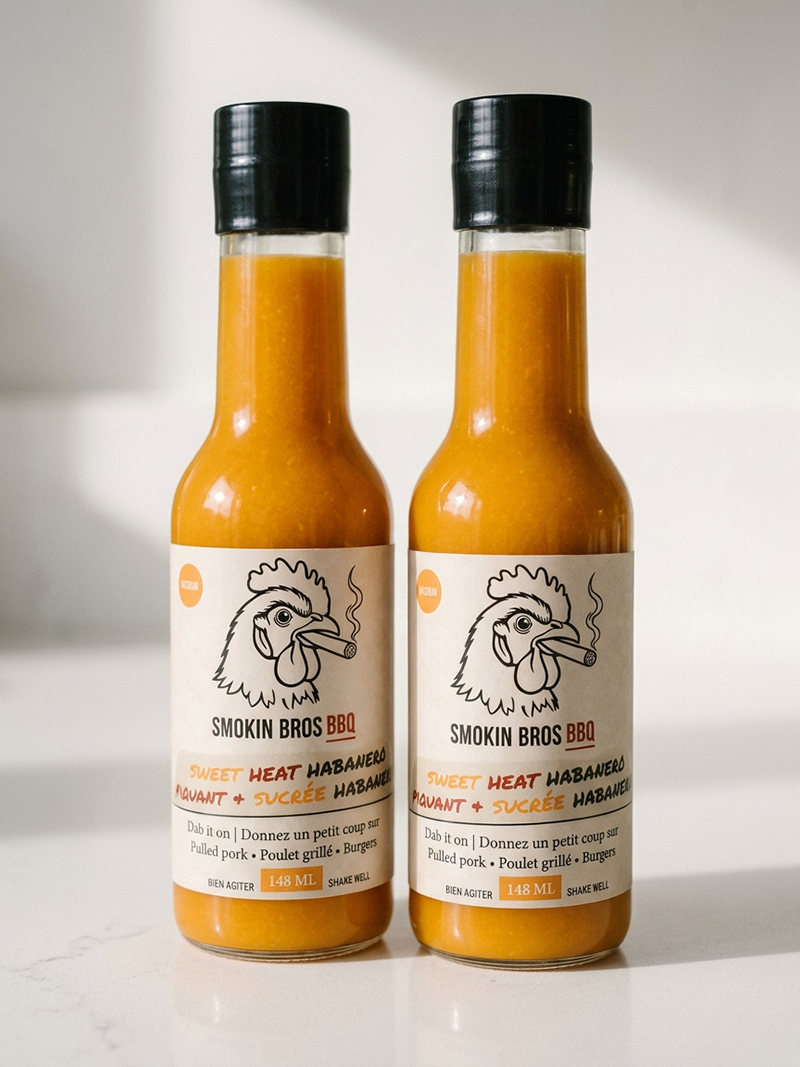Two bottles of Smokin Bros BBQ Sweet Heat Habanero sauce with orange sauce, cream labels, and black caps, standing on a white surface against a white background.