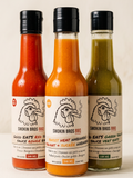 Three Smokin Bros BBQ sauce bottles standing side by side against a light beige background. The left bottle contains red sauce, the middle bottle contains orange sauce, and the right bottle contains green sauce.